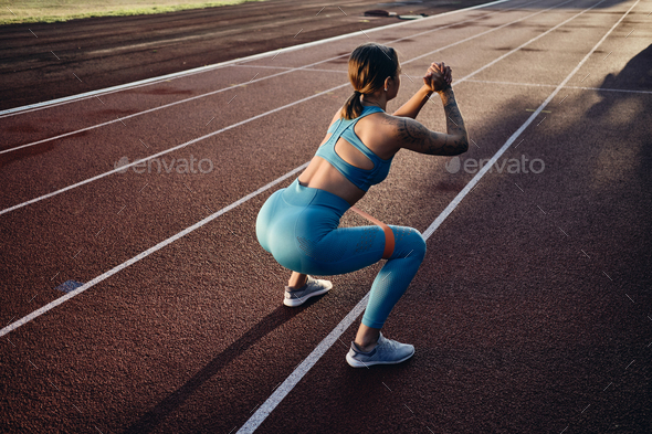 Attractive athlete girl in stylish sportswear squatting with rubber band during training on stadium