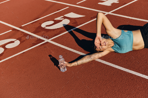 Tired athlete girl in sportswear lying on runner track dreamily covering eyes with hand from sun