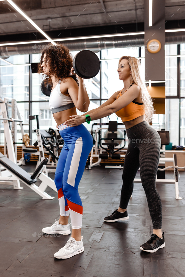 Slender girl in stylish bright sports clothes doing back squats with barbell and another athletic