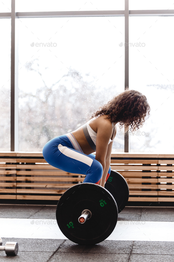 Slim curly dark-haired girl dressed in sports clothes is doing back squats with heavy barbell in the