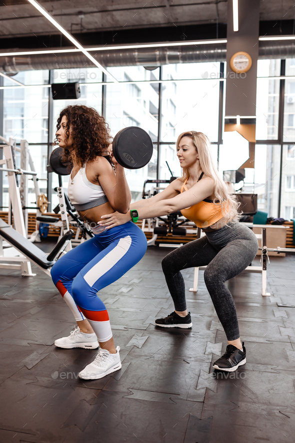 Slender girl in stylish bright sports clothes doing back squats with barbell and another athletic