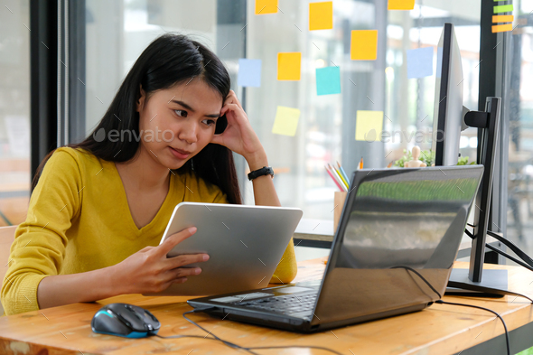 Asian female programmer wear yellow shirts, look at laptop screen and hold a tablet.