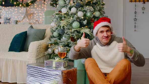 Young Man in Santa Hat Sitting on Floor Surrounded By Gift Boxes and Christmas Tree alt