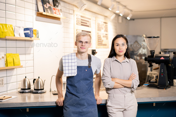 Two young workers of contemporary cafe or restaurant standing by workplace