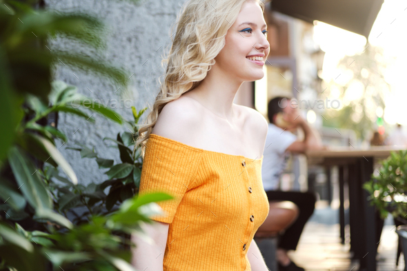 Side view of beautiful cheerful blond girl happily looking away in courtyard of cafe