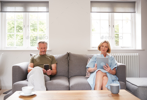 Anti-Social Senior Couple Sitting On Sofa At Home Using Digital Tablet And Mobile Phone