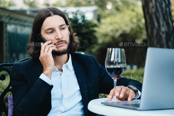 Latin businessman with glass of wine thoughtfully talking on cellphone working with laptop in cafe - Stock Photo - Images