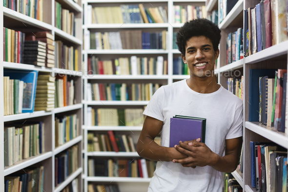 Cheerful african american guy staying between bookshelves, holding book ...