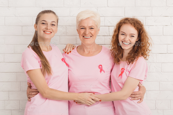 Three Women In Breast Cancer T-Shirts Embracing Against White Wall