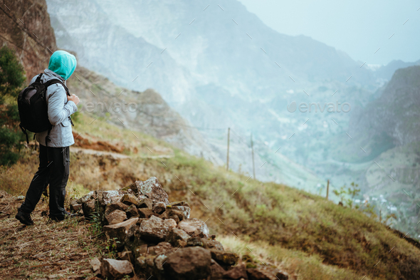 Traveler with backpack staying on the edge of path leading through rural landscape with mountains - Stock Photo - Images