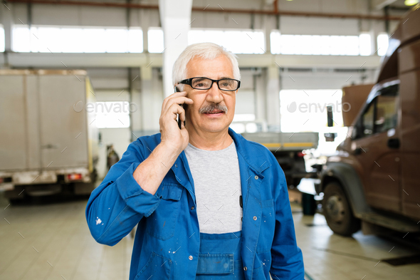 Senior technician in workwear and eyeglasses consulting client on mobile phone