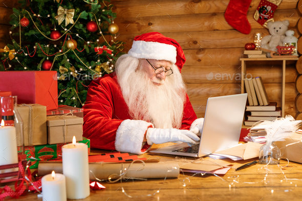 Santa Claus sitting by laptop while looking through xmas wishes in e-mail