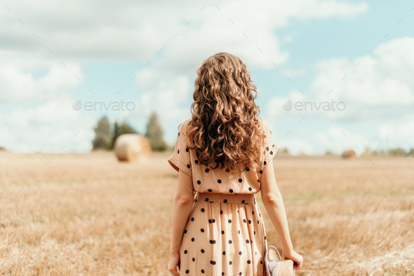 Woman in beige polka dot dress with curly hair, straw hat standing on harvested field with straw