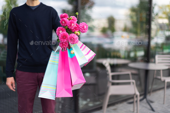 Romantic man giving flowers bouquet and bunch of pink gift bags near cafe. Ready for birthday party - Stock Photo - Images