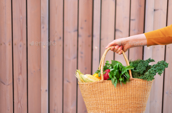 Woman hand holding straw basket with organic vegetables over wooden background. Healthy food - Stock Photo - Images