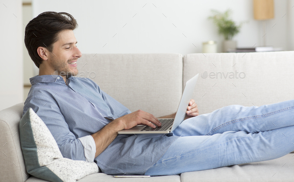 Guy Working On Laptop Computer Lying On Sofa At Home
