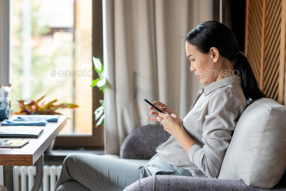 Young casual woman with smartphone sitting on couch in restaurant and messaging