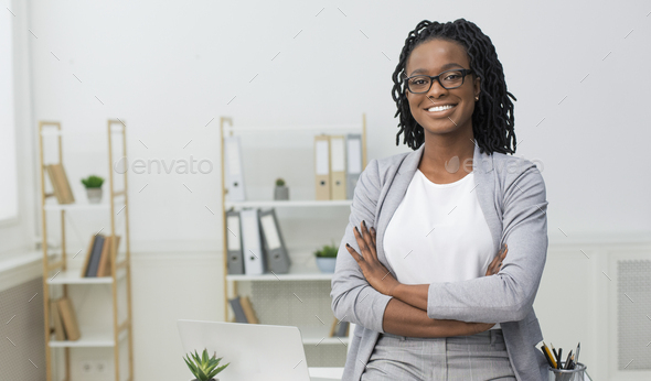 Smiling Black Businesswoman Sitting On Desk In Modern Office
