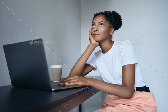 Attractive African American woman dreamily working on laptop with coffee in co-working space