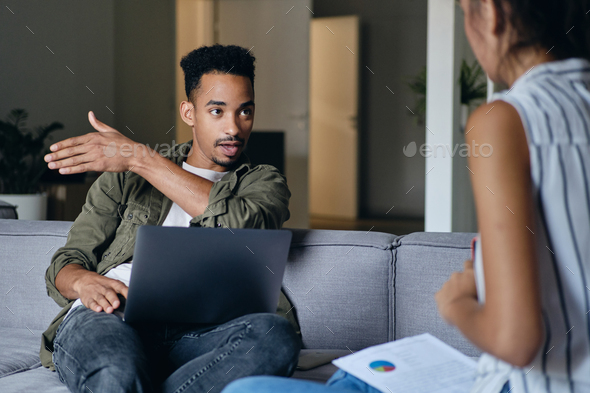 African American man with laptop emotionally discussing work with colleague in co-working space