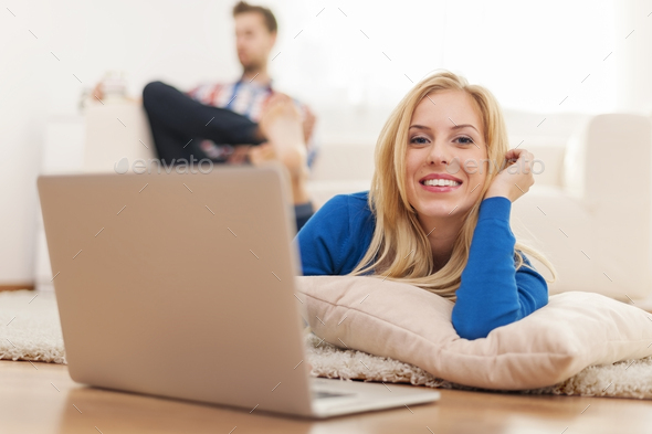 Beautiful blonde woman lying down on carpet at home and using laptop