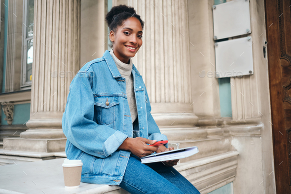 African American student girl in denim jacket with cellphone and book happily looking in camera - Stock Photo - Images