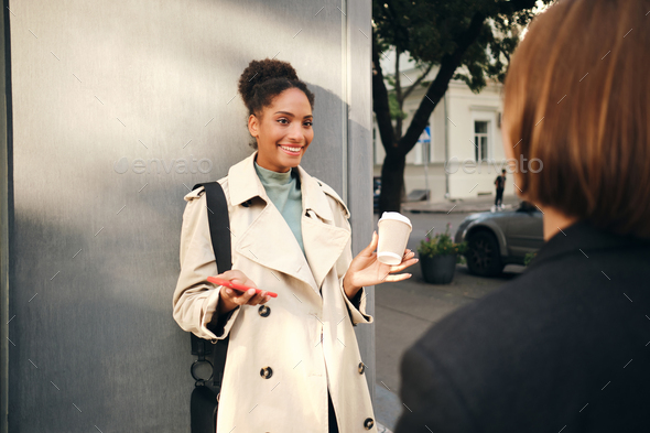 Beautiful African American girl in stylish trench coat emotionally talking  with friend outdoor Stock Photo by garetsworkshop