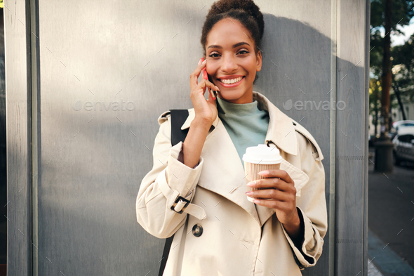 Happy African American girl in trench coat joyfully looking in camera talking on cellphone - Stock Photo - Images