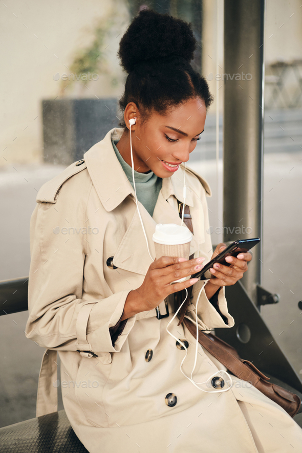 African American girl in stylish trench coat and earphones happily using cellphone at bus stop - Stock Photo - Images