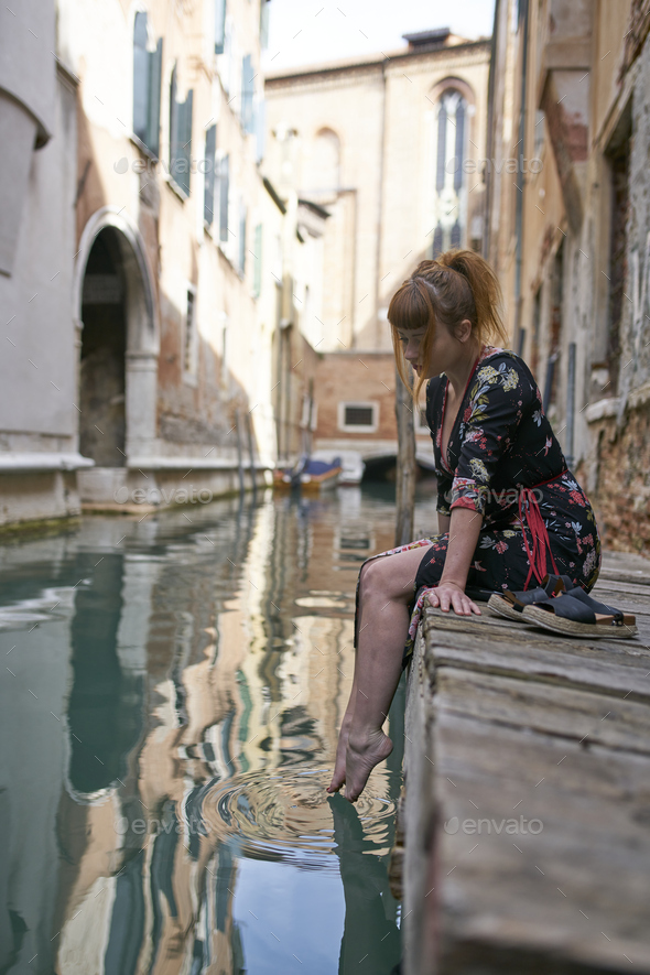 Caucasian redhead woman with floral dress sitting near a canal in Venice barefoot - Stock Photo - Images