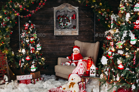Little Santa boy in red suite sitting in big arm chair near Christmas tree - Stock Photo - Images