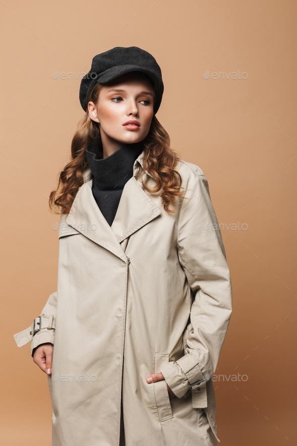 Beautiful girl with wavy hair in trench coat and cap holding hands in pockets over beige background - Stock Photo - Images