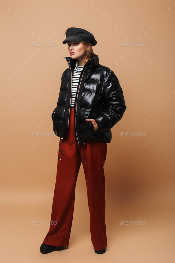 Stylish model with wavy hair in cap, wide pants and black down jacket posing over beige background