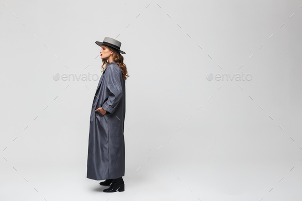 Young woman in hat and cloak holding hand in pocket looking aside over gray background - Stock Photo - Images