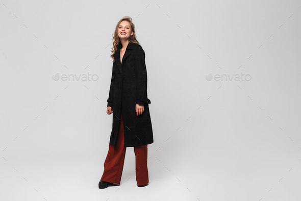 Stylish smiling woman in black coat and red wide trousers joyfully looking in camera at studio - Stock Photo - Images