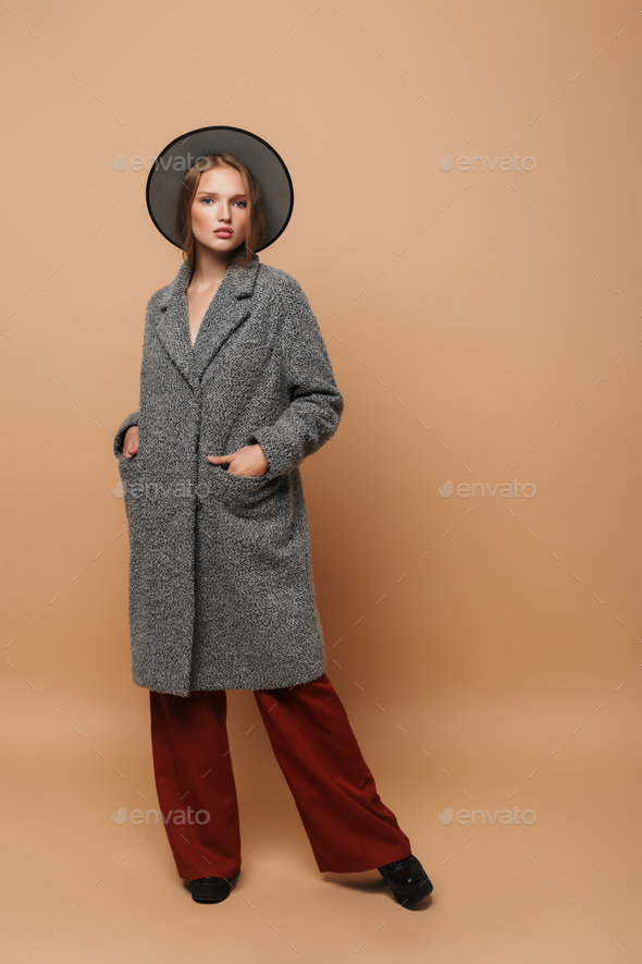 Stylish model with hat in gray coat and wide pants posing over beige background