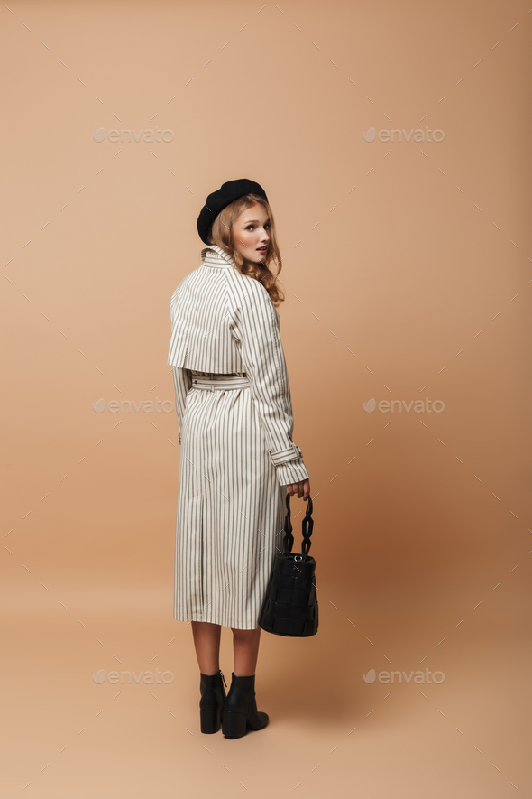 Young attractive woman in coat and black hat holding bag thoughtfully looking in camera from back - Stock Photo - Images