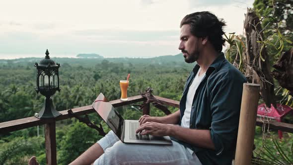 A young, handsome man with a beard works on a laptop in a cafe overlooking an incredible sunset. alt