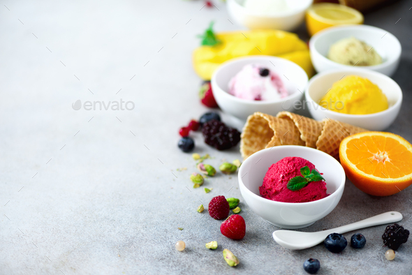Ice cream balls in bowls, waffle cones, berries, orange, mango, pistachio on grey concrete - Stock Photo - Images