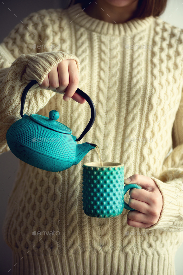 Woman in warm knitted woolen sweater holds turquoise teapot and pouring herbal tea into handmade cup - Stock Photo - Images