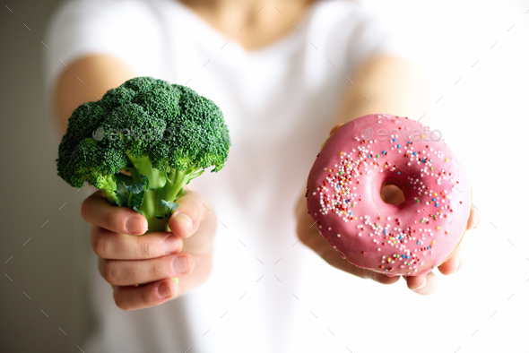 Young woman in white T-shirt choosing between broccoli or junk food, donut. Healthy clean detox - Stock Photo - Images