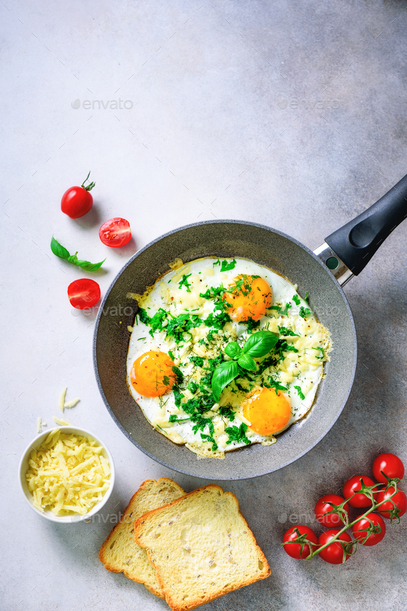 Fried eggs in pan, herbs, spices, cherry tomatoes, cheese toasts, basil for breakfast on grey