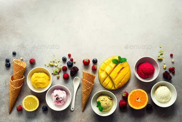Ice cream balls in bowls, waffle cones, berries, orange, mango, pistachio on grey concrete - Stock Photo - Images