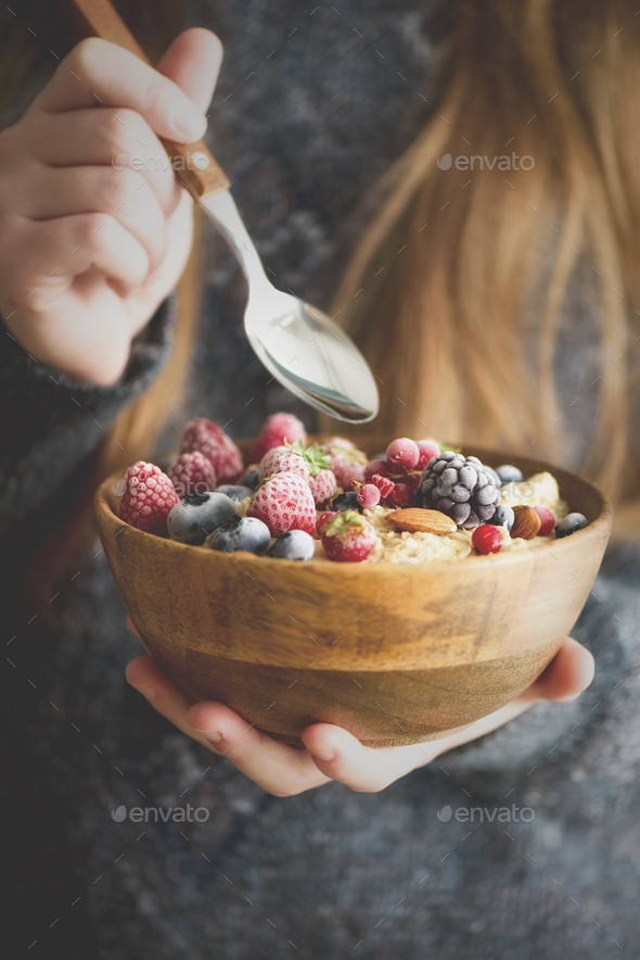 Girl hands holding oatmeal porridge with frozen berries, almonds in wooden bowl. Healthy breakfast - Stock Photo - Images