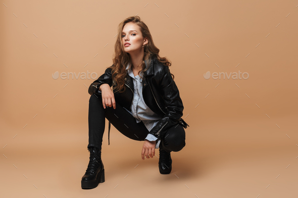 Young gorgeous woman with wavy hair in black leather jacket and shirt sitting down - Stock Photo - Images