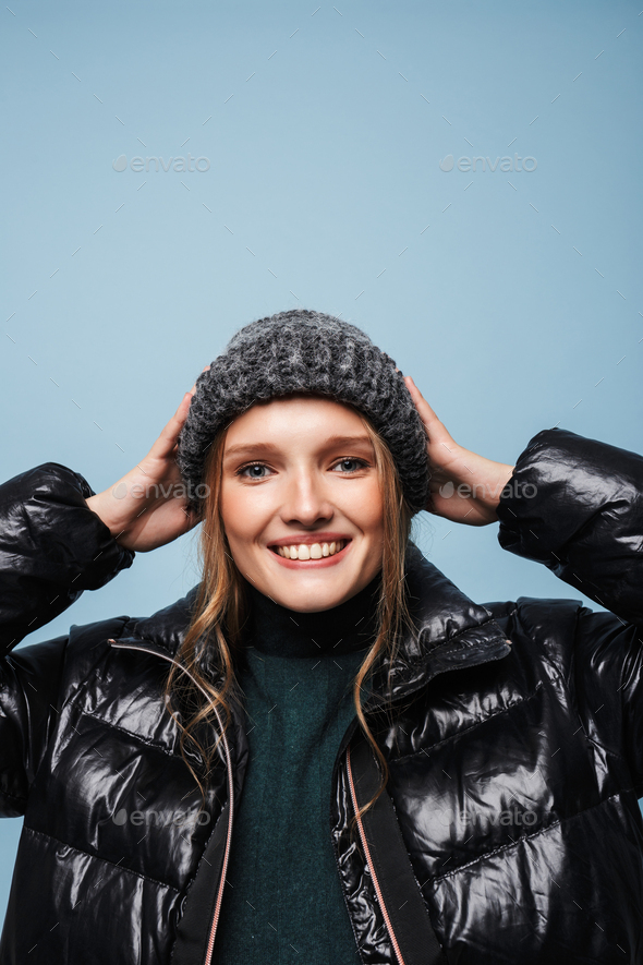 Young beautiful smiling woman in black down jacket and knitted hat joyfully looking in camera