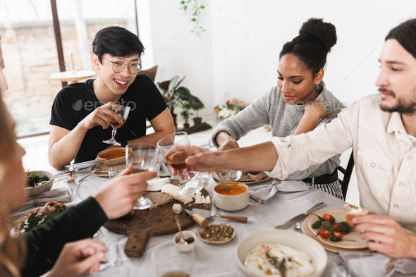 Group of attractive international friends sitting at the table full of food happily eating together - Stock Photo - Images