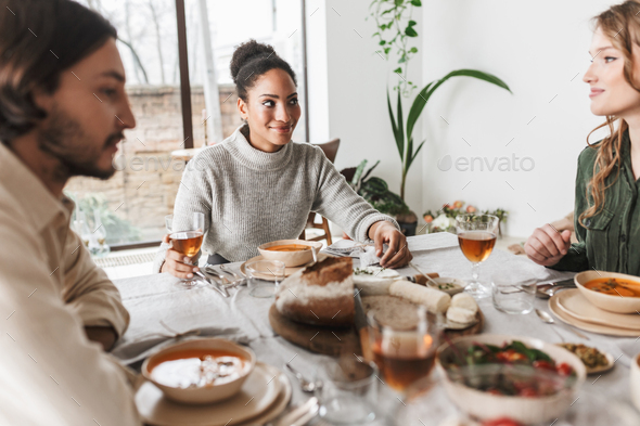 Pretty smiling african american woman sitting at the table full of food dreamily looking friend - Stock Photo - Images
