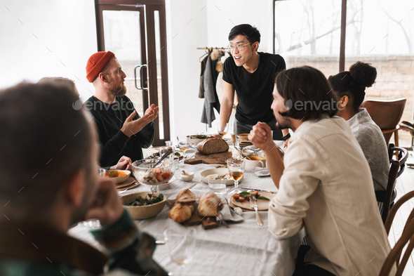 Young asian man in eyeglasses and black T-shirt leaning on table happily talking with colleagues - Stock Photo - Images