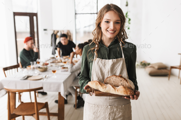 Young cheerful woman with wavy hair in white apron holding bread on board in hands - Stock Photo - Images